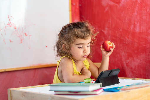 Two and half years old preschooler girl learning how to hold pen and write. She is wearing a yellow dress. Background is red painted wall. She is holding a red apple. Shot indoor with a full frame mirrorless camera.