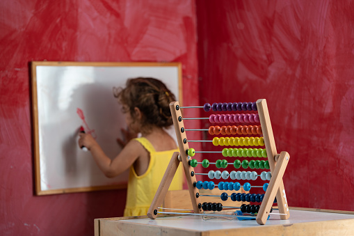 Two and half years old preschooler girl learning how to hold pen and write. She is wearing a yellow dress. Background is red painted wall. A multicolored abacus is seen on desk. Shot indoor with a full frame mirrorless camera.