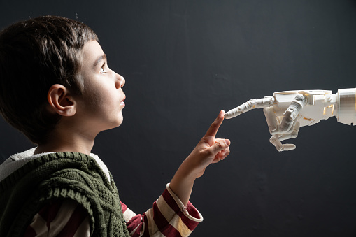 Six years old elementary schoolboy meeting humanoid robot. He is touching robotic hand's finger. The background is black. Shot with a full frame mirrorless camera.