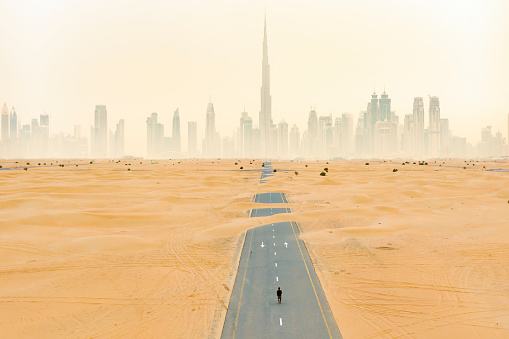 View from above, stunning aerial view of an unidentified person walking on a deserted road covered by sand dunes with the Dubai Skyline in the background. Dubai, United Arab Emirates.