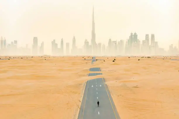 Photo of View from above, stunning aerial view of an unidentified person walking on a deserted road covered by sand dunes with the Dubai Skyline in the background. Dubai, United Arab Emirates.