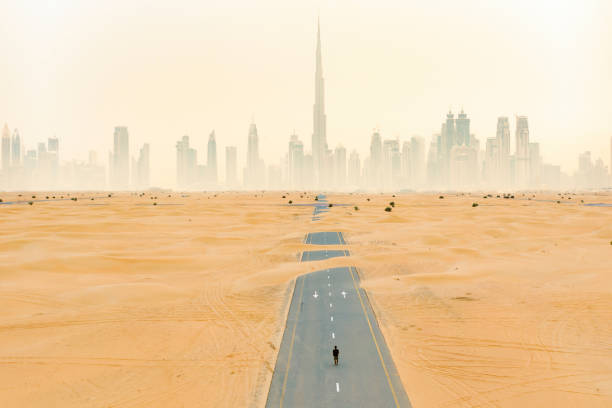 view from above, stunning aerial view of an unidentified person walking on a deserted road covered by sand dunes with the dubai skyline in the background. dubai, united arab emirates. - fog desert arabia sunset imagens e fotografias de stock