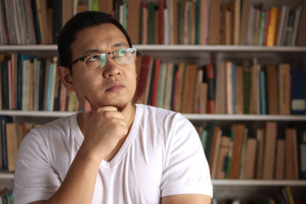 istock Asian man thinking while standing in front of bookshelf, education concept, studying in library, serious expression 1280408748