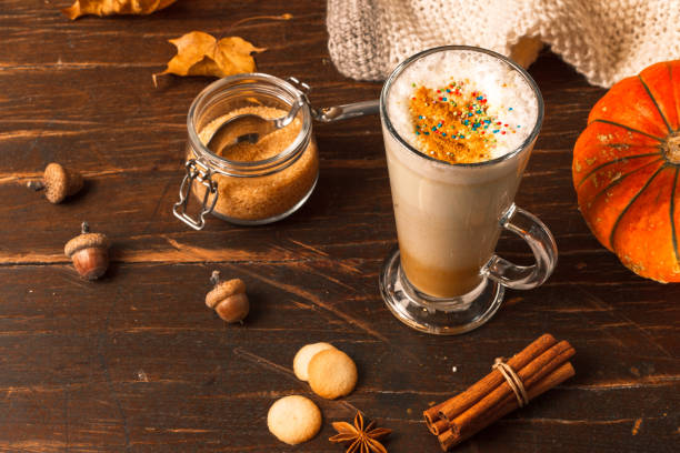 Latte in a tall spiced glass on a dark wooden table with dried maple leaves, pumpkin and cinnamon sticks and cookies stock photo