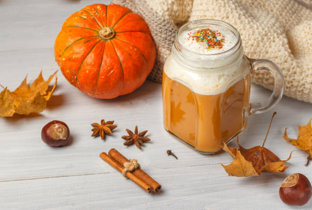 pumpkin latte with milk foam in a glass mug, a knitted blanket and an orange pumpkin on a light wooden table. Horizontal, with space ,close-up stock photo