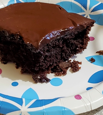 Homemade corner piece of single layer chocolate cake with chocolate icing on a paper birthday plate