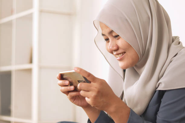 istock Beautiful Asian muslim woman using phone while relaxing on sofa, playing games or watching streaming online video, happy smiling expression 1280423524