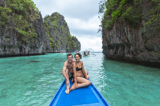 A mixed race couple on a romantic tropical vacation smile directly at the camera while sitting on the bow of a boat in their swimsuits. They are traveling in the Philippines and rocky island cliffs rise out of the water on both sides of the boat.