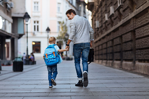 Young blond boy with a backpack walking to school with his dad