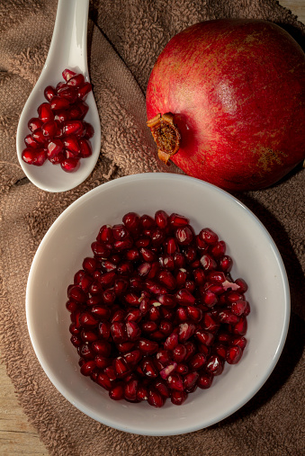Aerial close up view of grains of pomegranate and fruit in a bowl and a white tasting spoon on the wooden table and brown cloth.  Natural vitamin supplement for a healthy diet