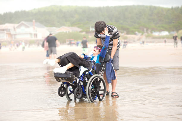 Young biracial Asian Caucasian man pushing disabled little brother in wheelchair on beach