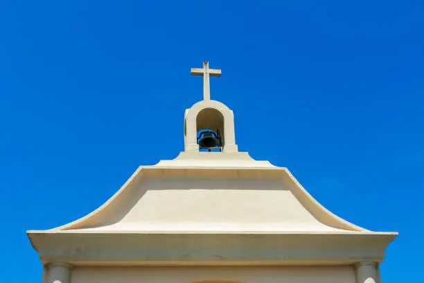 Exterior view of a top of small Catholic church with bell tower, one bell and cross under birght blue sky.