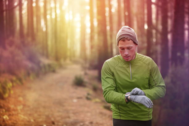 Trail running runner heart rate monitor watch Trail running runner looking at heart rate monitor watch running in forest wearing warm jacket sportswear, hat and gloves. Male jogger running training in woods. stopwatch photos stock pictures, royalty-free photos & images