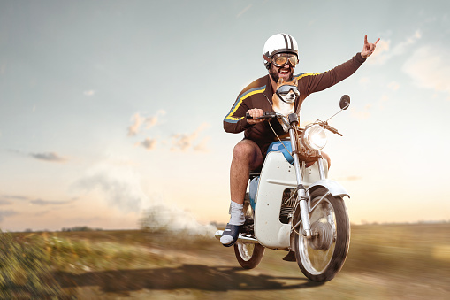 A man with a vintage helmet and goggles is riding a retro style moped. His left hand is raised forming devil horns. His pet dog is placed inside his jacket also wearing goggles. Motion blurred background with copy space.