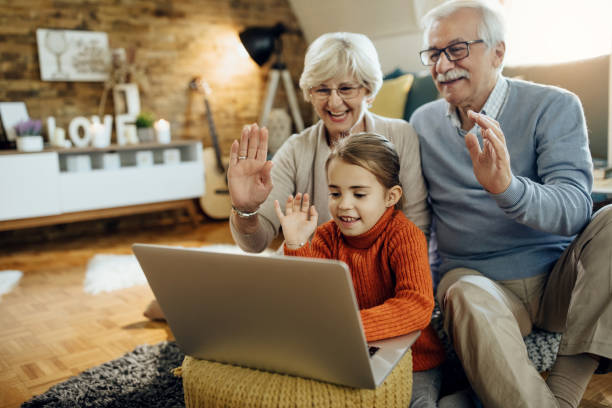 Happy little girl and her grandparents greeting someone while having video chat on laptop at home.