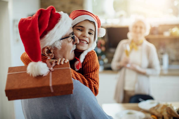 Grateful little girl embracing her grandfather while receiving Christmas present from him. Joyful granddaughter and grandfather embracing after exchanging gifts on Christmas day at home. multi generation family christmas stock pictures, royalty-free photos & images