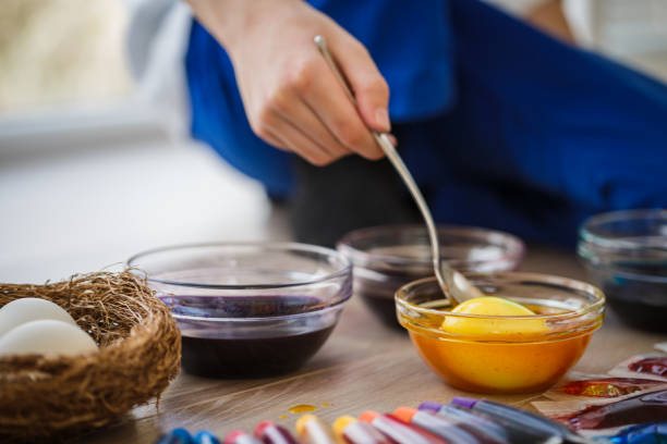 Close up of glass bowls with colorful artificial inks where a kid is painting a yellow Easter egg to welcome holiday season.