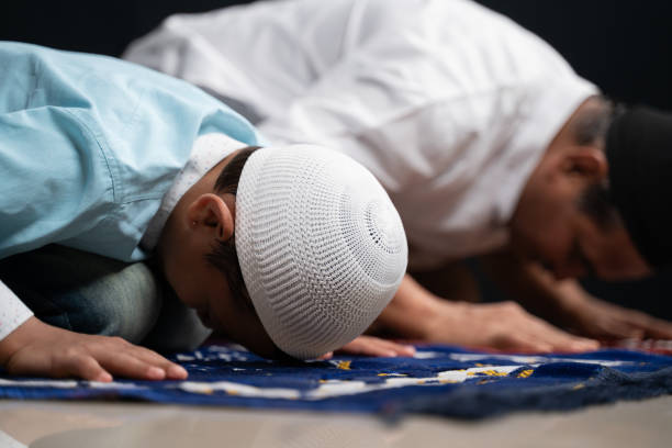Muslim father and son praying or performing Salah while sitting on Prayer rug and touching head to mihrab or mosque printed on rug. Muslim father and son praying or performing Salah while sitting on Prayer rug and touching head to mihrab or mosque printed on rug father allah stock pictures, royalty-free photos & images