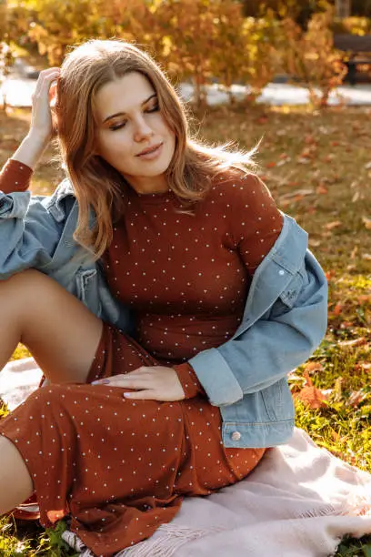 Photo of A young woman is sitting on the autumn ground in a polka-dot dress and jeans.