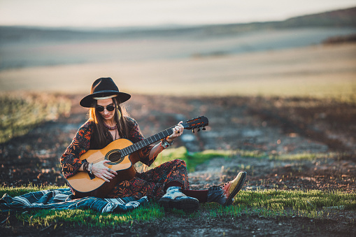 Hippie woman with guitar playing music and relaxing on field in sunny day.