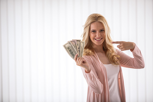 Copyspace, waist up of satisfied adult woman holding a bunch of money in one hand and pointing at it with her index finger.