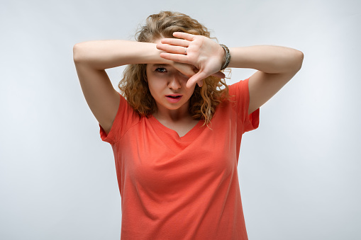 Portrait of frightened young woman with curly hair in casual t shirt covering her eyes with hands. Human emotions, facial expression concept. Studio shot, white background, isolated