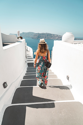 Caucasian woman tourist in long dress with straw hat walking down a staircase in Oia town on a hot summer day. Traditional white houses and streets of Santorini.