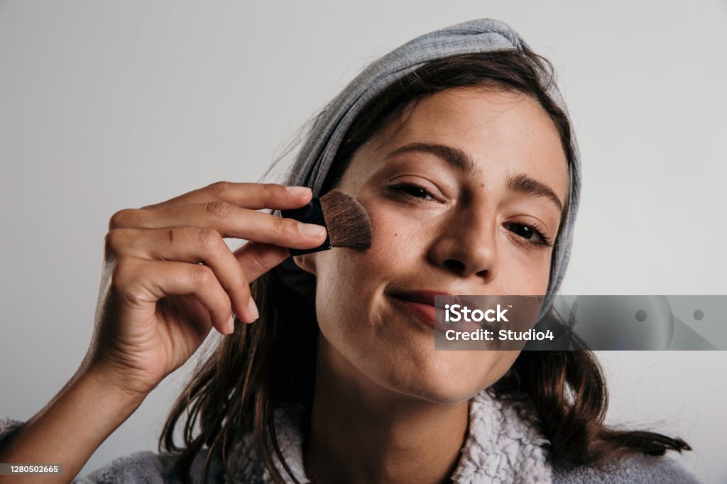 A little blush for a nicer glow A photo of a beautiful young adult woman having her time, looking at the camera with a joyful face wearing a light gray hairband, putting the blush gently on her cheeks. Isolated in a studio. Adult Stock Photo