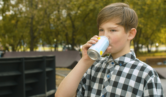 Portrait of teenager boy drinking soda water near pump track. Refreshing drink, rest after training. City park, summer day