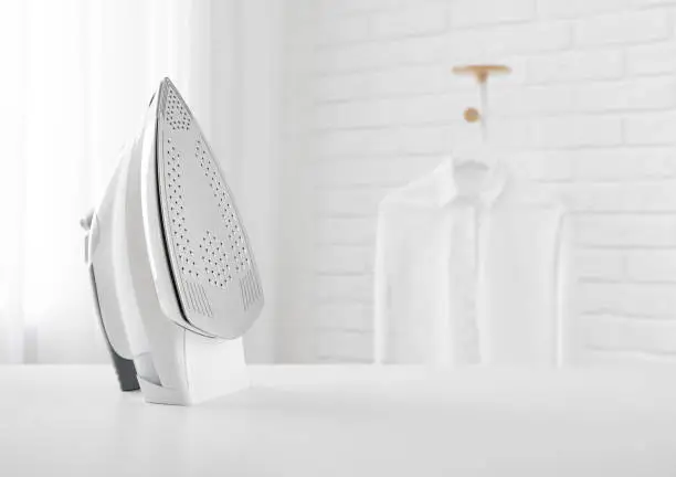 Photo of Electric iron on table in blurred room with clothes rack