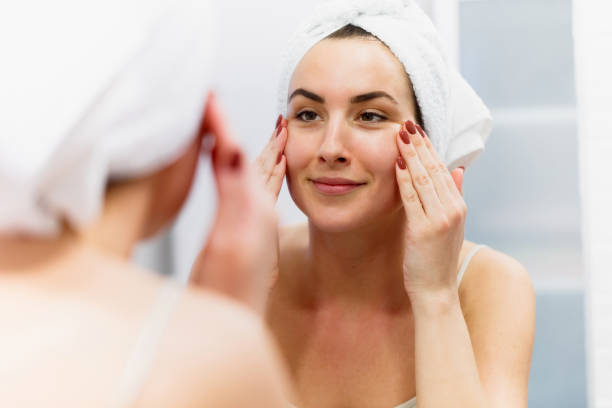 濡れた髪にタオルを着た笑顔の女性が鏡の前で肌を引き締める - human face rubbing women beauty treatment ストックフォトと画像