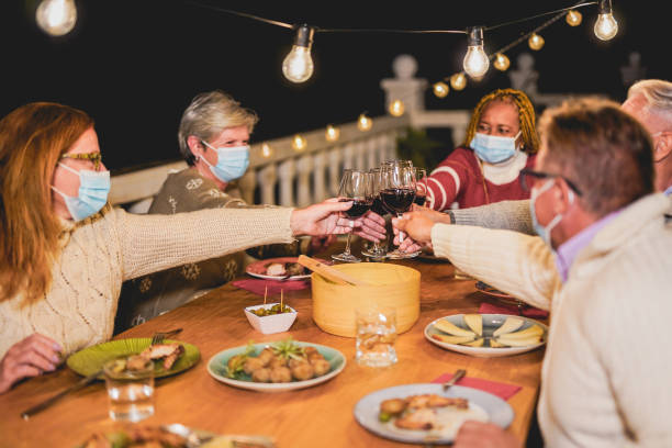 happy multiracial senior people cheering with red wine at dinner while wearing surgical face mask for coronavirus - dinner friends christmas imagens e fotografias de stock