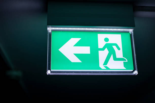 istock Green and white exit sign hangs on the ceiling of the corridor, lighted in the darkness. 1280543690