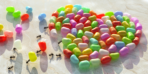 A conceptual image of ants working together in harmony arranging jelly beans in the shape of a heart on a marble counter / worktop. Three ants are carrying the last beans to complete the shape as other ants move around.