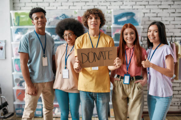 istock Group of cheerful diverse young volunteers smiling at camera while standing in charitable organization office, Guy holding card with Donate lettering 1280557947