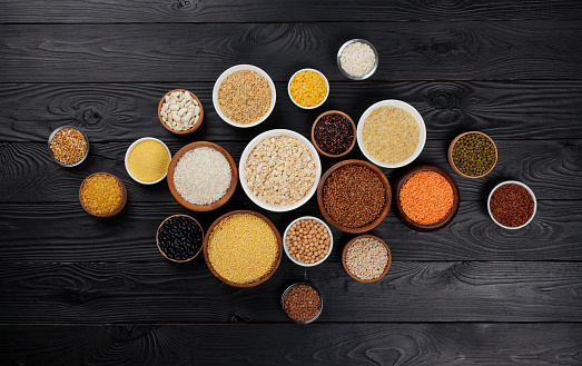 Different cereals, grains, seeds, groats, legumes and beans in bowls, top view of raw porridge collection on black wooden background with copy space