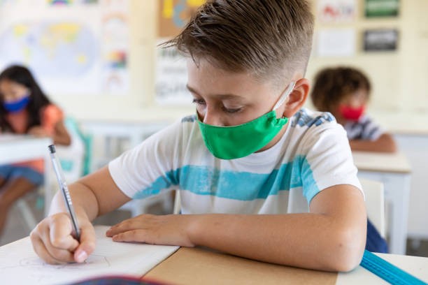 istock Boy wearing face mask writing while sitting on his desk at school 1280563016