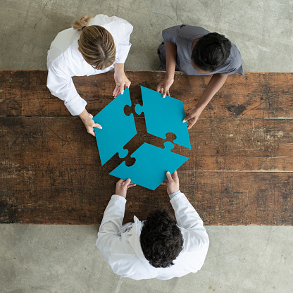 Three people dressed in medical clothing stand around a table and each hold a puzzle piece which together form a whole. They are pictured from the aerial view.