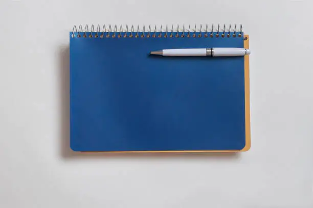 Blue spiral notebook with white pen attached on gray surface. Flat view.