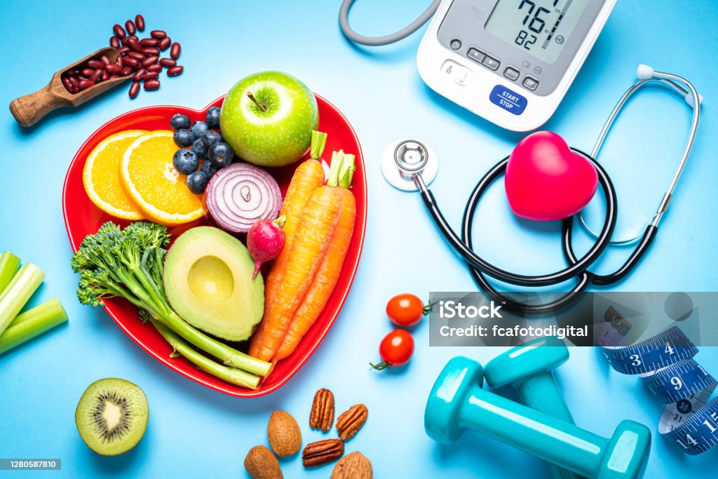 Healthy eating, exercising, weight and blood pressure control Healthy lifestyle concepts: red heart shape plate with fresh organic fruits and vegetables shot on blue background. A digital blood pressure monitor, doctor stethoscope, dumbbells and tape measure are beside the plate  This type of foods are rich in antioxidants and flavonoids that prevents heart diseases, lower cholesterol and help to keep a well balanced diet. High resolution 42Mp studio digital capture taken with SONY A7rII and Zeiss Batis 40mm F2.0 CF lens Healthy Lifestyle Stock Photo