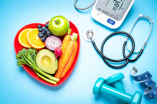 Healthy lifestyle concepts: red heart shape plate with fresh organic fruits and vegetables shot on blue background. A digital blood pressure monitor, doctor stethoscope, dumbbells and tape measure are beside the plate.  This type of foods are rich in antioxidants and flavonoids that prevents heart diseases, lower cholesterol and help to keep a well balanced diet. High resolution 42Mp studio digital capture taken with SONY A7rII and Zeiss Batis 40mm F2.0 CF lens