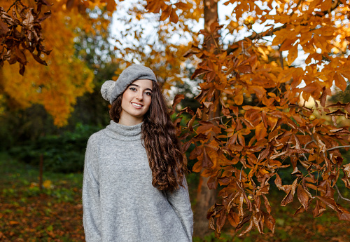 Attractive stylish smiling girl with curly hair walking in park dressed in warm grey autumn trendy fashion, wearing beret hat. Space for text.