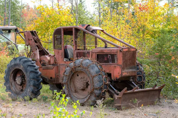 An old, rusty, abandoned heavy tractor with a plow on the front. The tractor is next to a woods. Chains on front tires. No identifying markings.