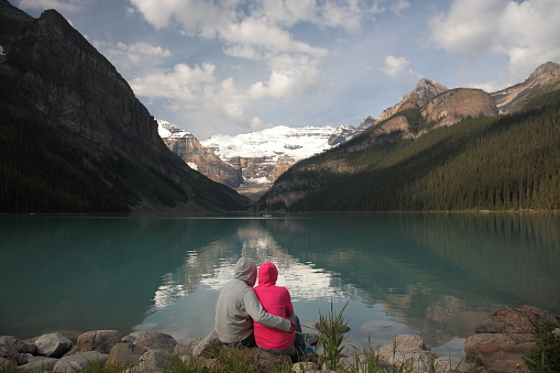 Couple siting on rock by the lake watching Mount Victoria with Victoria Glacier and Lake Louise during summer in Banff National Park, Canadian Rockies, Alberta, Canada.