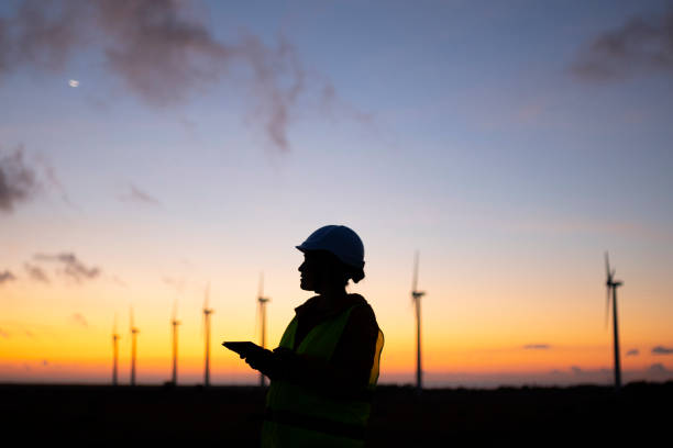 Renewable Energy Systems. Electricity Maintenance Engineer working on the field at a Wind Turbine Power station at dusk. Electrical engineer working for the energy industry, supervising the condition of the Electrical Power Equipment in a wind turbines farm power station at night. Checking the data and the results of measurements with digital tablet. Technology and Global Business. carbon neutrality photos stock pictures, royalty-free photos & images
