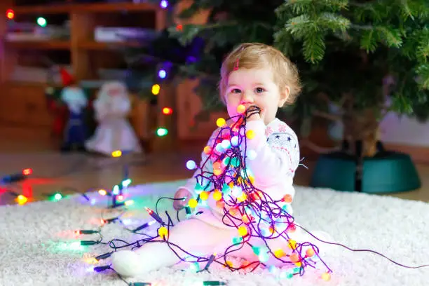 Adorable baby girl holding colorful lights garland in cute hands. Little child in festive clothes decorating Christmas tree with family. First celebration of traditional holiday called Weihnachten.