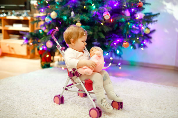 Cute adorable baby girl having fun with with doll carriage. Beautiful toddler child sitting in stroller with toy at home. Happy daughter. With Christmas tree and lights. Cute adorable baby girl having fun with with doll carriage. Beautiful toddler child sitting in stroller with toy at home. Happy daughter. With Christmas tree and lights buggy eyes stock pictures, royalty-free photos & images