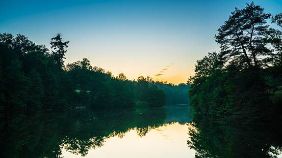 Germany, Stuttgart, Magical orange sunset sky behind tree and forest silhouette reflecting in water of lake baerensee