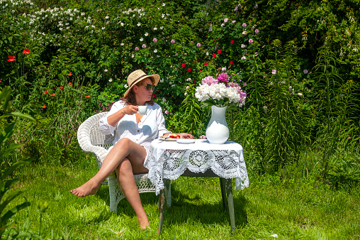 Smiling woman in the garden drinking tea or drinking coffee while sitting at a table with flowers in a vase