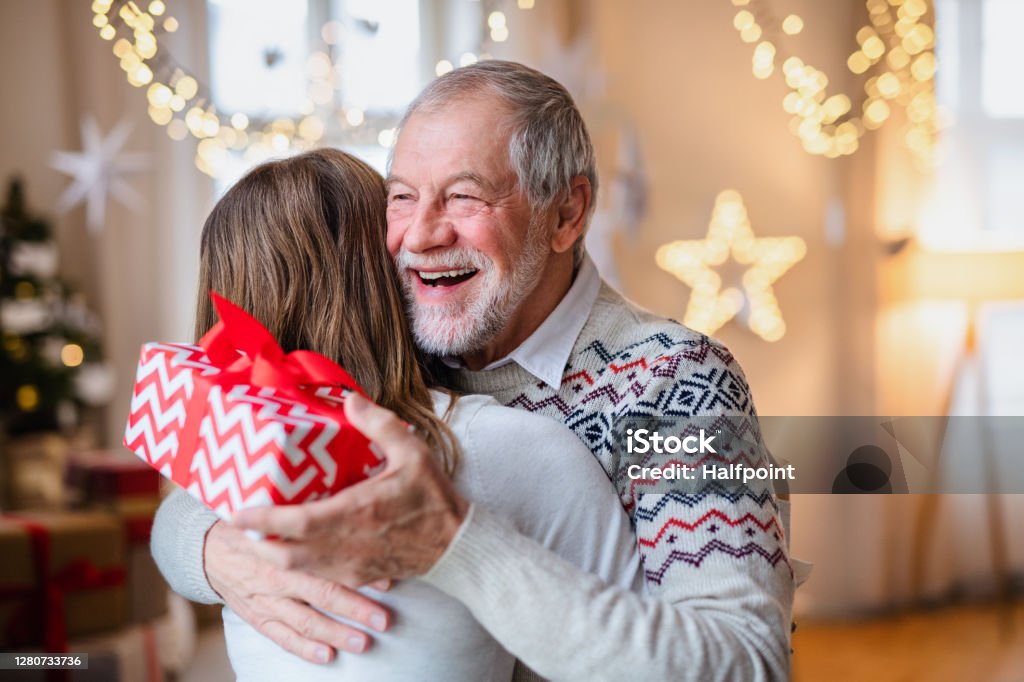 Young woman giving present to happy grandfather indoors at home at Christmas. Portrait of young woman giving present to happy grandfather indoors at home at Christmas. Christmas Stock Photo